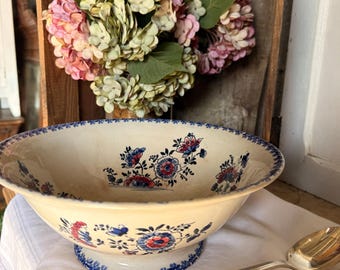May include: A cream-colored ceramic bowl with a blue floral pattern and a blue rim. The bowl sits on a pedestal base with a matching blue floral design. Two silver spoons are next to the bowl. A bouquet of pink and green flowers is in the background.