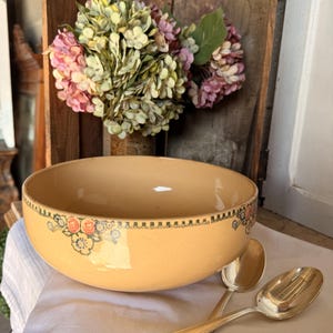 May include: A large, tan ceramic bowl with a floral design around the rim and a pair of silver spoons. The bowl is set on a white cloth, with a bouquet of flowers in the background.
