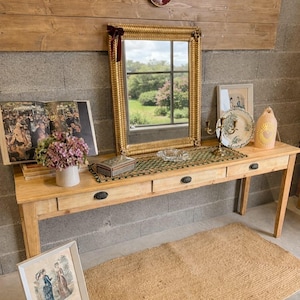 Peut inclure: Une console en bois clair à trois tiroirs, ornée d'un miroir doré, d'œuvres d'art et d'objets décoratifs. Un tapis tissé et une estampe encadrée reposent sur le sol. Le mur derrière la table est en bois et en béton.
