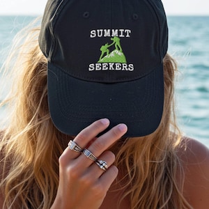 Puede incluir: Gorra de béisbol negra con el texto "SUMMIT SEEKERS" en blanco y un gráfico de montaña verde. La gorra la lleva una persona con pelo largo y rubio, con el mar de fondo.