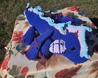 Puede incluir: Un dragón de ganchillo hecho a mano en tonos de azul, morado y rosa. El dragón presenta un cuerpo texturizado, detalles rizados y ojos de seguridad negros. Está sobre una mesa cubierta con un mantel con temática otoñal.