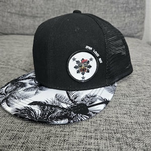 May include: Black and white snapback hat with a palm tree design on the brim. The hat features a mesh back and a circular patch with text that reads "SPAM LOVES RICE".