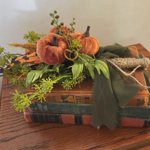 May include: A stack of three vintage books topped with two small orange velvet pumpkins, green artificial foliage, dried grasses, and a dark olive green fabric ribbon tied around the collection with a piece of natural twine.
