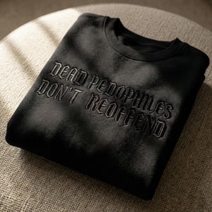 May include: Black sweatshirt with the embroidered text "DEAD PEDOPHILES DON'T REOFFEND" in a gothic font. The sweatshirt is folded and resting on a neutral-toned surface, with soft lighting creating shadows.