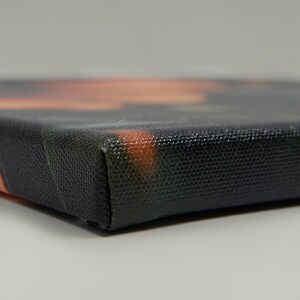 Peut inclure: Gros plan sur une impression sur toile tendue. La toile a une surface noire textur&eacute;e avec un coin mis au point. Les bords montrent des touches de couleurs orange et verte, sugg&eacute;rant une &oelig;uvre d'art paysag&egrave;re ou abstraite.