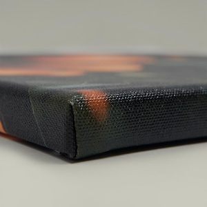 May include: Close-up of a stretched canvas print. The canvas has a black textured surface with a corner in focus. The artwork on the canvas is blurred, with orange and green hues visible.