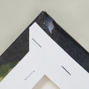 May include: Close-up of a canvas print corner. The canvas is stretched over a wooden frame and secured with staples. The canvas has a black textured surface and a white backing. A clear plastic corner protector is visible.