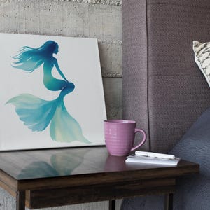 May include: A canvas art print featuring a mermaid silhouette in shades of blue and teal. The artwork is displayed on a small wooden side table, next to a pink mug, a pen, and a small notebook. The background includes a textured wall and a bed with a patterned pillow.