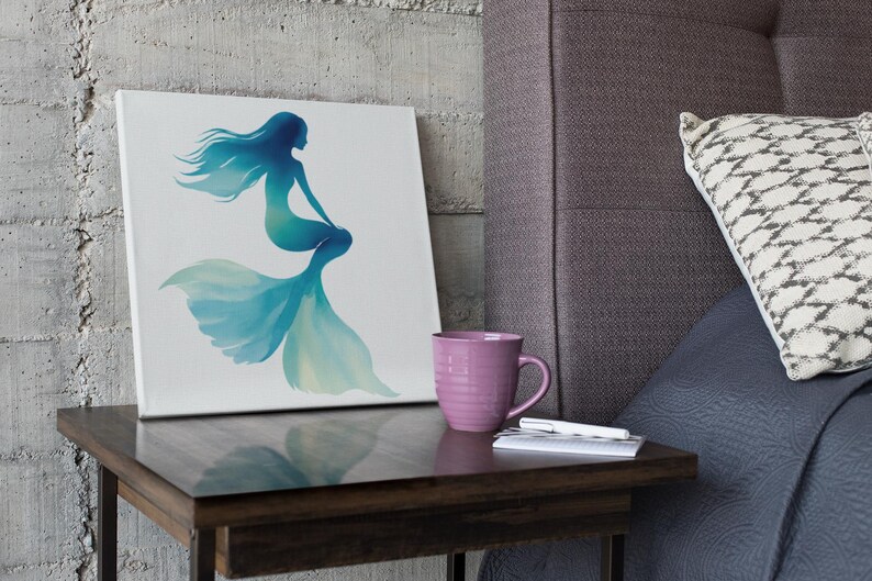 May include: A canvas art print featuring a mermaid silhouette in shades of blue and teal. The artwork is displayed on a small wooden side table, next to a pink mug, a pen, and a small notebook. The background includes a textured wall and a bed with a patterned pillow.