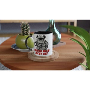 Könnte beinhalten: Weißer Keramikbecher mit schwarzem Innenraum und Henkel. Der Becher zeigt eine Cartoon-Bulldogge im Bademantel, die eine Tasse hält, mit dem Text "SPRICH MICH NICHT AN!" in Rot. Der Becher steht auf einem Holzuntersetzer.