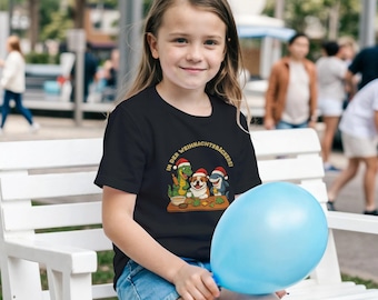May include: A young girl wearing a black t-shirt with a festive graphic. The shirt features a cartoon scene of a dinosaur, dog, and shark wearing Santa hats and baking. The text "IN DER WEIHNACHTSBÄCKEREI" is above the characters. She is holding a blue balloon.