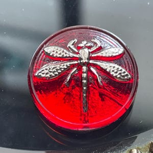 May include: A round, red button with a silver dragonfly design. The dragonfly has detailed wings and body, set against a vibrant red background. The button is likely made of glass or a similar material, and is a decorative element.