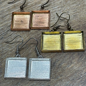 May include: Four pairs of square-shaped earrings with different metal finishes: copper, gold, and silver. Each earring has a small, square frame with a hook for the ear. The earrings are displayed on a wooden surface.
