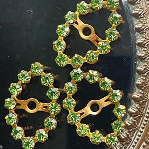 May include: Three vintage gold-toned metal buckles, each with a circular design. The circles are adorned with small, round, light green rhinestones. The buckles have a central bar with a hole for attachment, set against a black background.
