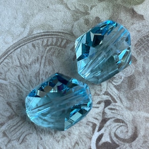 May include: Two faceted, light blue crystal beads. The beads have multiple cut surfaces that reflect light, creating a sparkling effect. The beads are set against a patterned, neutral-toned background.