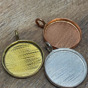 May include: Three round pendant trays in gold, copper, and silver tones. Each tray has a loop for a chain and a textured surface for adhering a design. The trays are displayed on a wooden surface.