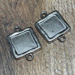 Puede incluir: Dos engastes cuadrados plateados para la fabricación de joyas. Cada engaste tiene un área empotrada para incrustar una piedra o diseño, con pequeños bucles en cada lado para la fijación. Los engastes se muestran sobre una superficie de madera.