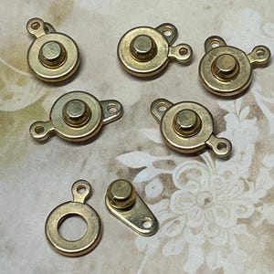 May include: A collection of seven small, gold-colored metal clasps. The clasps have a circular or rectangular base with a raised, rounded center and a small loop for attachment. The clasps are designed for use in jewelry making or crafts.