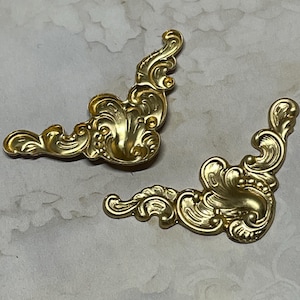 May include: Two ornate, gold-colored corner brackets with a detailed, swirling design. The decorative hardware pieces are made of metal and are ideal for furniture restoration or craft projects.