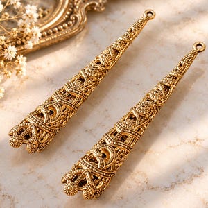 May include: Two elongated, cone-shaped gold-toned metal pendants featuring intricate filigree cutouts, floral motifs, and textured geometric patterns. Each pendant has a small circular loop at the top for attachment, showcasing a detailed, openwork design.