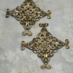 May include: Two ornate, gold-colored filigree embellishments. Each piece features a symmetrical, cross-like design with intricate scrollwork and floral details. The embellishments are set against a patterned, off-white background.