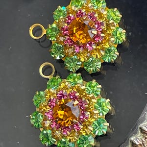 May include: Two floral-shaped earrings with a gold-tone setting. Each earring features a large, amber-colored center stone surrounded by pink, yellow, and green crystals. The earrings have a loop at the top for attachment.