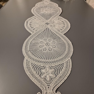 Könnte beinhalten: Dekorativer, cremefarbener Tischläufer mit einem aufwendigen, durchbrochenen Design. Der Läufer weist ein sich wiederholendes Muster aus floralen und geometrischen Formen mit einem gewellten Rand auf. Der Läufer ist ca. 152 cm lang.