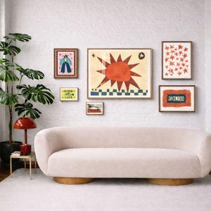 Peut inclure: Un salon avec un mur de briques blanches, pr&eacute;sentant une galerie d'impressions d'art encadr&eacute;es. Un canap&eacute; incurv&eacute; rose clair repose sur un tapis blanc. Une lampe rouge et une plante en pot sont &agrave; gauche. L'art comprend un motif de soleil et le mot "SHINDIG".
