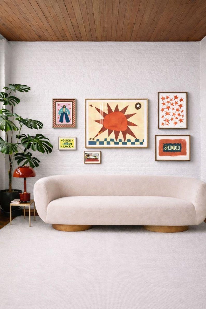 Peut inclure: Un salon avec un mur de briques blanches, pr&eacute;sentant une galerie d'impressions d'art encadr&eacute;es. Un canap&eacute; incurv&eacute; rose clair repose sur un tapis blanc. Une lampe rouge et une plante en pot sont &agrave; gauche. L'art comprend un motif de soleil et le mot "SHINDIG".