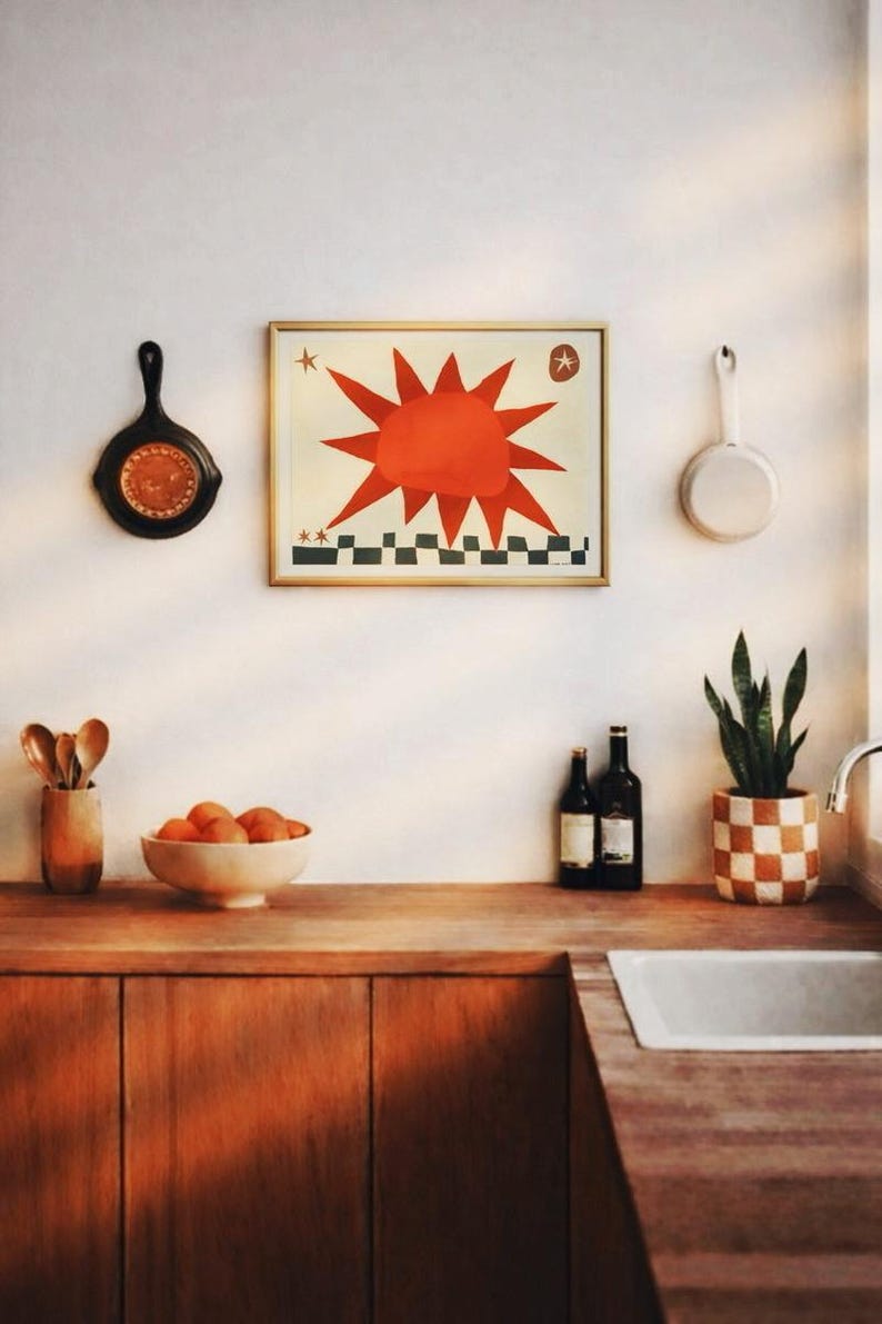 Peut inclure: Int&eacute;rieur de cuisine avec plan de travail et placards en bois. Une &oelig;uvre d'art encadr&eacute;e avec un soleil rouge est accroch&eacute;e au mur. Il y a un bol d'oranges, des bouteilles de vin et un pot de plante &agrave; carreaux.