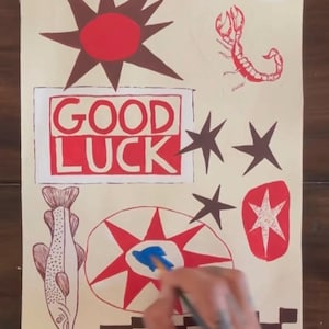 Puede incluir: Una hoja de papel rectangular con ilustraciones pintadas a mano en rojo, marr&oacute;n y azul. El dise&ntilde;o incluye un sol rojo, un escorpi&oacute;n, un pez, estrellas y un recuadro rojo con el texto GOOD LUCK. Se ve un pincel aplicando pintura azul.