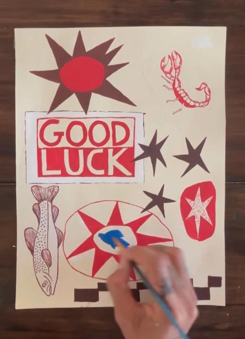 Puede incluir: Una hoja de papel rectangular con ilustraciones pintadas a mano en rojo, marr&oacute;n y azul. El dise&ntilde;o incluye un sol rojo, un escorpi&oacute;n, un pez, estrellas y un recuadro rojo con el texto GOOD LUCK. Se ve un pincel aplicando pintura azul.