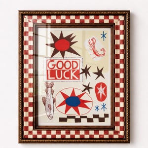 Puede incluir: Una l&aacute;mina de arte enmarcada con fondo crema e ilustraciones gr&aacute;ficas en rojo, marr&oacute;n y azul que incluyen un pez, un escorpi&oacute;n, estrellas y un sol. El texto GOOD LUCK est&aacute; impreso en rojo. El marco tiene un borde a cuadros rojos y blancos.