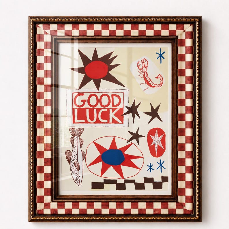 Puede incluir: Una l&aacute;mina de arte enmarcada con fondo crema e ilustraciones gr&aacute;ficas en rojo, marr&oacute;n y azul que incluyen un pez, un escorpi&oacute;n, estrellas y un sol. El texto GOOD LUCK est&aacute; impreso en rojo. El marco tiene un borde a cuadros rojos y blancos.