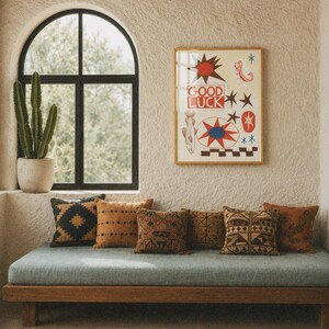 Puede incluir: Una l&aacute;mina de arte mural enmarcada con el texto GOOD LUCK en rojo, rodeado de ilustraciones de un sol, un escorpi&oacute;n, un pez y formas geom&eacute;tricas en rojo, azul y negro sobre un fondo crema, expuesta sobre un banco de madera con cojines.