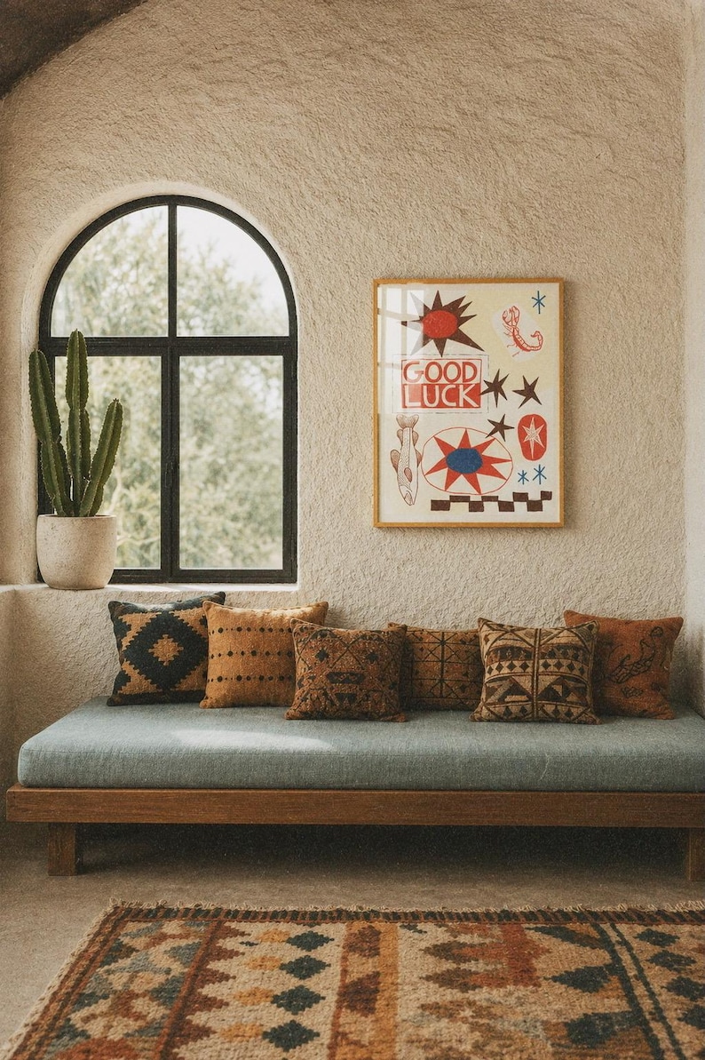 Puede incluir: Una l&aacute;mina de arte mural enmarcada con el texto GOOD LUCK en rojo, rodeado de ilustraciones de un sol, un escorpi&oacute;n, un pez y formas geom&eacute;tricas en rojo, azul y negro sobre un fondo crema, expuesta sobre un banco de madera con cojines.