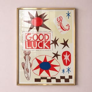 Puede incluir: Una l&aacute;mina art&iacute;stica enmarcada con fondo crema y el texto rojo GOOD LUCK dentro de un borde rectangular. El dise&ntilde;o incluye un escorpi&oacute;n rojo, un pez, un sol marr&oacute;n oscuro, una estrella roja y azul, y un patr&oacute;n de cuadros marr&oacute;n oscuro.