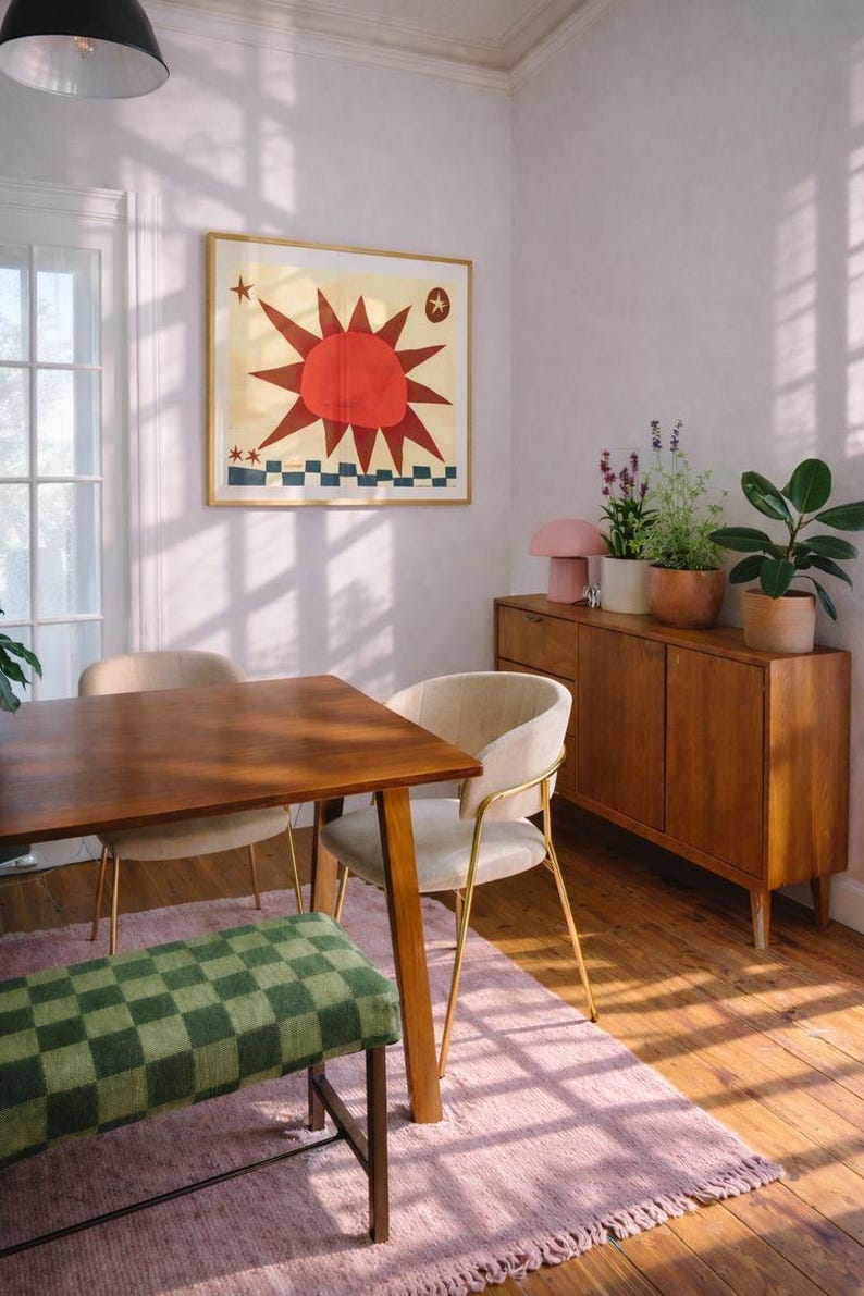 Peut inclure: Int&eacute;rieur de salle &agrave; manger avec une table en bois, des chaises et un banc &agrave; carreaux verts. Une &oelig;uvre d'art encadr&eacute;e avec un soleil rouge est accroch&eacute;e au mur. Un tapis rose et un meuble en bois avec des plantes et une lampe rose.