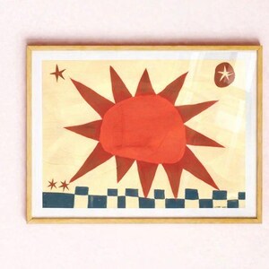 Peut inclure: Impression d'art encadr&eacute;e repr&eacute;sentant un soleil stylis&eacute; avec des rayons rouges et un cercle central rouge. L'&oelig;uvre comprend des &eacute;toiles et un motif &agrave; carreaux bleu et blanc en bas. L'impression est dans un cadre en bois.