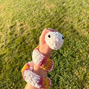 May include: A handmade crochet snake toy in white, pink, and yellow. The snake has a fluffy texture and black button eyes. The snake is coiled around a wrist, showcasing its design and colors.
