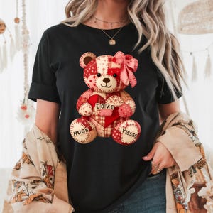 May include: Black t-shirt featuring a patchwork teddy bear design. The bear has a red heart with the word "LOVE" and the words "HUGS" and "VISSES" on its paws. The bear is adorned with a pink bow.