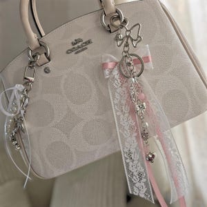 May include: A cream-coloured Coach handbag with a signature monogram pattern, featuring two silver-toned metal bag charms attached to the handles. The charms include white pearl beads, pink ribbons, white lace strips, and small silver-toned metal butterfly charms.