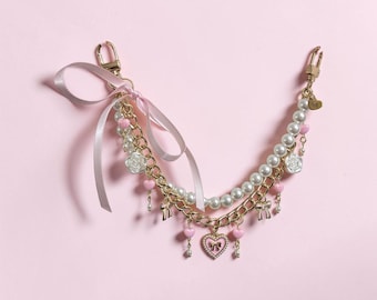 Puede incluir: Una correa de cadena dorada para bolso con un lazo de cinta de satén rosa claro, cuentas de perlas blancas, pequeños dijes de corazón rosa, dijes de rosas blancas y un colgante central en forma de corazón con un pequeño lazo, con dos mosquetones.