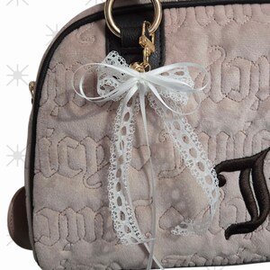 Puede incluir: Un bolso de mano rosa claro con asa negra y herrajes dorados. El bolso presenta un lazo de encaje blanco y una letra bordada marr&oacute;n. El bolso tiene un patr&oacute;n acolchado.