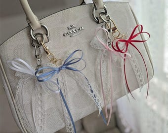 May include: A white Coach handbag featuring a textured surface and silver hardware. Two decorative bag charms are attached to the silver rings, each consisting of a metal clasp, a white lace ribbon, and a satin bow in blue, pink, or red.