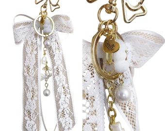 May include: A gold-toned metal keychain featuring a bow-shaped clasp, a large white lace ribbon, and a circular ring. Dangling charms include a small gold heart engraved with the text Cirelle, a white pearl bead, and a small white bear-shaped charm.