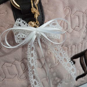 Puede incluir: Un lazo de encaje blanco con detalles de cinta de raso est&aacute; sujeto a un bolso. El bolso es de color rosa claro con una superficie texturizada y herrajes dorados. El lazo es un accesorio decorativo.