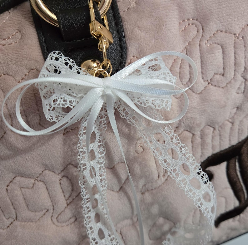 Puede incluir: Un lazo de encaje blanco con detalles de cinta de raso est&aacute; sujeto a un bolso. El bolso es de color rosa claro con una superficie texturizada y herrajes dorados. El lazo es un accesorio decorativo.