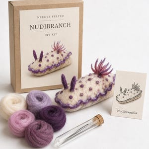 May include: A needle felted nudibranch DIY kit featuring a cardboard box labelled NEEDLE FELTED NUDIBRANCH DIY KIT, a finished felted nudibranch, five small balls of wool in cream and purple shades, a glass vial with needles, and a card with a drawing.