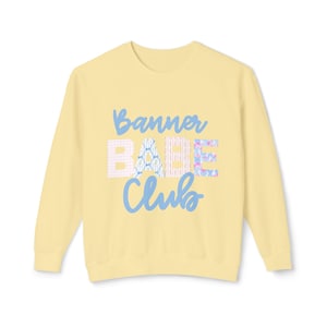 Może przedstawiać: Jasnożółta bluza z długimi rękawami. Z przodu widnieje napis "Banner Babe Club" w niebieskim skrypcie i wzorzystych literach, w tym kratka i kwiatowe wzory. Bluza wykonana jest z miękkiego materiału.